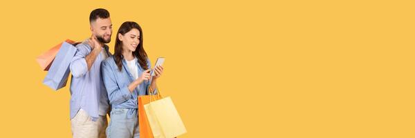 Happy spouses holding shopper bags and using smartphone, couple browsing mobile application with sale offers and discounts, standing over yellow background photo