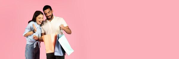 Loving handsome young hispanic man make surprise, presenting showing his girlfriend colorful shopping bags. Excited pretty millennial lady checking purchases, exclaiming and gesturing on pink photo