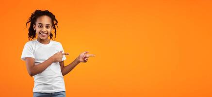 Look Here, Cool Offer. Portrait Of Smiling African American Girl Pointing With Two Fingers Aside At Free Copy Space On Yellow Wall, Happy Kid Presenting Empty Place For Your Design, Banner, Panorama photo