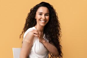 Portrait Of Smiling Young Woman Showing Arm After Coronavirus Vaccination, Happy Brunette Female Had Covid Vaccine Injection, Sitting On Chair With Rolled Up Sleeve Over Yellow Background, Copy Space photo