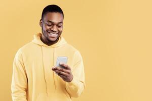 Cheerful African American Guy Using Cellphone Texting Reading Positive Message Standing Over Yellow Studio Background. Black Man Browsing Internet On Smartphone. New Mobile Application Concept photo