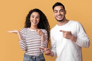 Check This. Cheerful Middle Eastern Couple Pointing At Open Palm, Happy Arab Man And Woman Demonstrating Invisible Object, Showing Copy Space While Standing Isolated On Yellow Background, Panorama photo