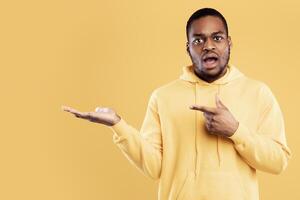 Look At This. Shocked Black Man Pointing Finger Showing Copy Space And Holding Invisible Object On Hand Posing Wearing Hoodie Over Yellow Studio Background. Panorama photo