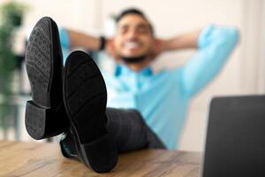 Young Arab employee relaxing after successful work, putting legs on table at office, selective focus. Millennial man resting at workplace with hands behind his head, copy space photo