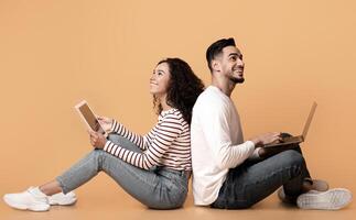 Online Entertainment. Happy Middle Eastern Couple With Laptop And Digital Tablet Sitting Back To Back And Looking Up, Cheerful Arab Spouses Using Modern Gadgets, Relaxing On Yellow Studio Background photo