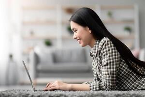 Side view of asian young woman using laptop at home, laying on carpet in living room, typing on computer keyboard, chatting with lover or friends while resting at weekend, copy space photo