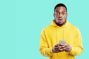 Shocked African American Guy Holding Phone Looking At Camera With Opened Mouth Posing Standing Over Blue Studio Background. New Mobile Application Concept photo