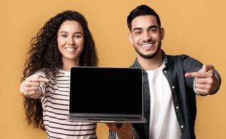 Great Website. Smiling middle eastern couple pointing at laptop with black screen for mockup, demonstrating copy space for your web design and advertisement, standing isolated over yellow background photo