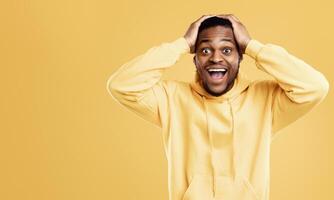 Excited Black Guy Shouting Touching Head Smiling Looking At Camera In Shock And Amazement Posing Standing On Yellow Studio Background, Wearing Bright Hoodie. Wow News, Excitement Concept photo