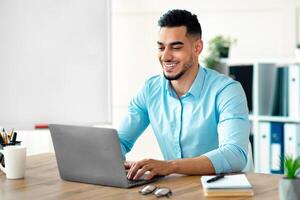 Handsome young Arab businessman working with laptop computer at his desk in office. Happy guy entrepreneur typing on pc keyboard, checking email, taking part in online meeting at workplace photo