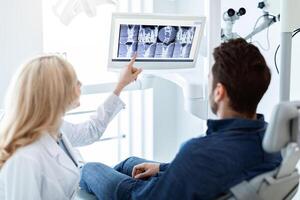 Modern dental clinic concept. Blonde woman dentist having conversation with patient in dental chair, poitning at LED display, showing results of treatment or explaining procedure photo