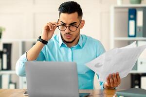 Stressed young Arab employee using laptop, holding documents, having problem with work task, making mistake in business project at office. Millennial guy having trouble meeting deadline photo