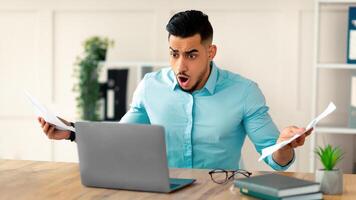 Shocked young Arab businessman with documents looking at laptop screen in dismay, shouting WTF, making mistake in project at office. Business crisis, error, deadline concept photo
