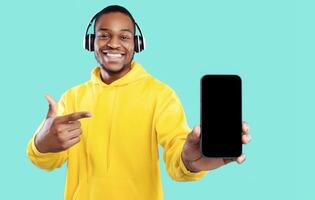 Music Application. Cheerful Black Man Showing Phone Blank Screen Wearing Wireless Headphones Smiling To Camera Posing Over Blue Studio Background. App Advertisement Mockup. Selective Focus photo