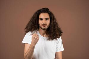 A young man with curly, long hair stands confidently against a brown background. He raises a fist, showing a powerful expression, embodying determination and strength. photo