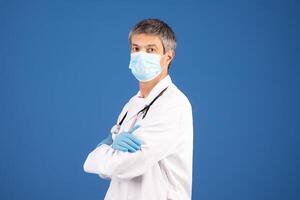 A mature Caucasian man in his 50s, dressed in a white coat, blue gloves, and a mask stands confidently against a solid blue background, representing a committed general physician. photo