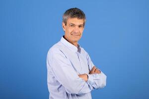 A mature Caucasian man in his 50s stands with arms crossed, smiling gently. He is dressed in a light blue shirt and is surrounded by a simple blue background, creating a relaxed atmosphere. photo
