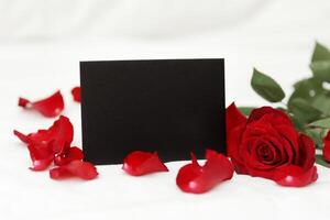 Black stylish invitation or greeting card with mockup and bright red rose flower and petals on white background. St. Valentines day celebration. Women's day. Copy space for text. Empty blank. photo