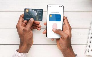 Online Payment And Biometric Identification Concept. Top Above View Of Black Man Holding Cellphone And Credit Card In Hand Showing Mobile Wallet App With Fingerprint Icon On Screen For Scanning photo