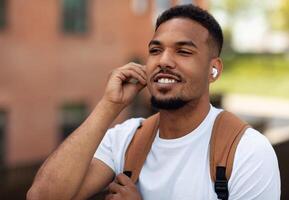 Closeup portrait of handsome black guy wearing earpods and listening to music outdoors, walking in urban city area, free space. Man resting after work and enjoying favorite song photo
