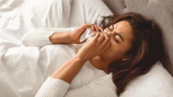 Sick black woman suffering from running stuffy nose and sore throat. Upset ill African American lady lying in bed, blowing her nose using paper napkin tissue, banner. Cold And Flu Concept photo