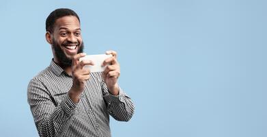 Gadget Addiction Concept. Portrait of happy black man playing mobile games on cellphone isolated over blue studio background, copy space. Satisfied male adult using his device, watching photo