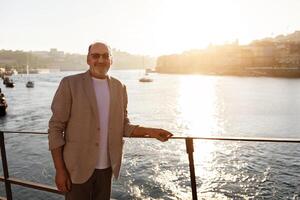 A man with glasses and a light jacket stands by a railing at the waterfront during sunset, looking relaxed as boats glide on the water, with a picturesque town illuminated in the background. photo