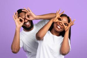 Having Fun. Portrait of joyful black couple smiling, looking at the camera through fingers, doing okay gesture and covering eyes, imitating eyeglasses. Facial expressions and emotions concept photo