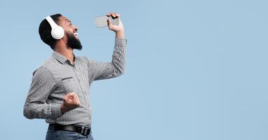 Full length photo of excited emotional black man singing his favorite song, using smartphone as microphone, studio shot