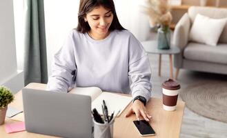 Business And Telecommuting Concept. High angle view of smiling young woman using her smartphone and modern laptop computer, reading text message, sitting at desk with gadgets, working at home office photo