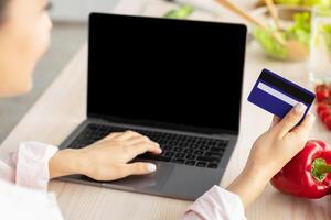 Easy Payment. Over shoulder view of smiling young woman using laptop sitting at dinner table, holding and showing debit credit card, selective focus, blurred background. Housewife ordering food photo
