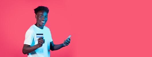 A young black man stands smiling, holding a smartphone in one hand and a credit card in the other. The bright pink backdrop enhances his joyful expression, highlighting engagement with technology. photo