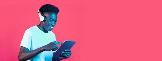 A young black man enjoys music through headphones while focused on his tablet. The dynamic pink background adds energy to the moment, highlighting his engagement and expression of joy. photo