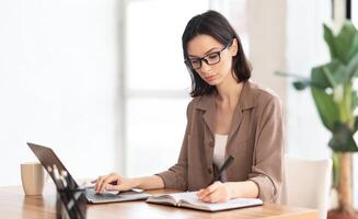 Note Taking. Portrait of female student using laptop and writing memo in her diary at modern office, copyspace photo