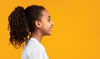 Side view profile portrait of cute African American girl with beaming smile and curly hair in ponytail looking aside at empty free copy space isolated on yellow studio background, banner, panorama photo