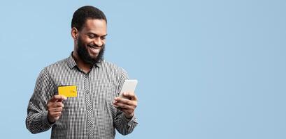Ecommerce Concept. Satisfied black guy holding credit card, using mobile phone isolated over blue studio background wall. Smiling african american man shopping, ordering via internet photo
