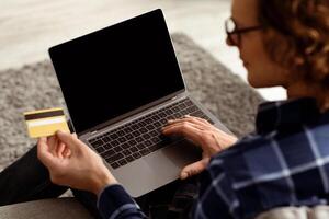 Back-View Of Man Using Laptop And Credit Card Shopping Online On Computer With Empty Screen Sitting At Home. Guy Purchasing And Paying In Internet Store. Cropped, Selective Focus, Mockup photo