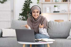Online Tutoring. Young muslim woman teacher having call with students, talking at laptop camera, sitting on couch at home photo