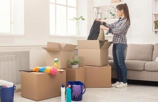New home cleaning. Young woman tidying up and unpacking boxes after moving to new apartment. Girl with various detergents, rags and mops in living room full of carton boxes. photo