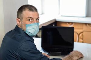 Coronacrisis Concept. Man Sitting At Laptop With Word COVID-19 On Screen, Wearing Medical Mask Working At Home. Selective Focus photo