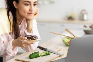 Interesting Reality Show Concept. Portrait of concerned millennial housewife watching film or movie in kitchen at home, looking at laptop computer screen while cooking, holding donut on plate photo
