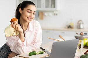 Interesting Reality Show. Portrait of smiling millennial housewife watching film or movie in kitchen at home, looking at laptop screen while cooking salad. Cheerful woman eating muffin cake photo