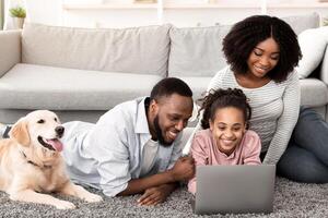 Portrait of happy loving black family lying on floor carpet with labrador, using laptop and watching or movie, browsing internet, spending time together at home in living room photo