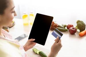 Ordering Food Online. Over the shoulder view of smiling woman using tablet with blank screen and holding credit card, ordering food delivery online, cooking salad in kitchen, free copy space photo
