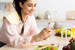 People And Technology Concept. Cheerful smiling millennial woman using mobile phone, standing in kitchen and leaning on counter, cheking social media or searching for new recipe, free copy space photo