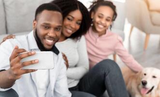 Beautiful African American family embracing and taking selfie with dog on mobile phone while resting at home and sitting on the floor, selective focus on gadget, blurred background. Technology photo