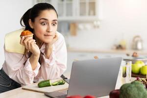 Exciting Movie Concept. Portrait of terrified Amazed surprised young lady watching film or reality show in kitchen at home, using laptop computer while cooking, eating muffin cake, leaning on counter photo