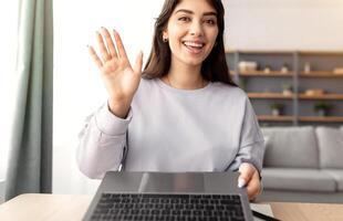 Online Call. Portrait of smiling young woman having virtual conference with friends, family or colleagues using computer, waving hello to webcam, sitting at desk at home office, view from screen photo