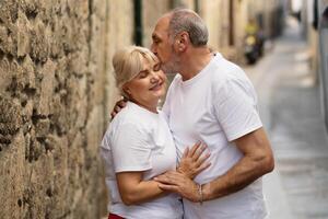 Couple stands closely in a narrow stone alley, smiling joyfully as one kisses the other's forehead. The soft summer light enhances their affectionate moment surrounded by rustic charm. photo