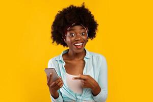 Excited African American Woman Pointing Finger At Smartphone Recommending Mobile Application For Your Phone Posing Over Pink Studio Background, Smiling To Camera. Check This Out Concept photo